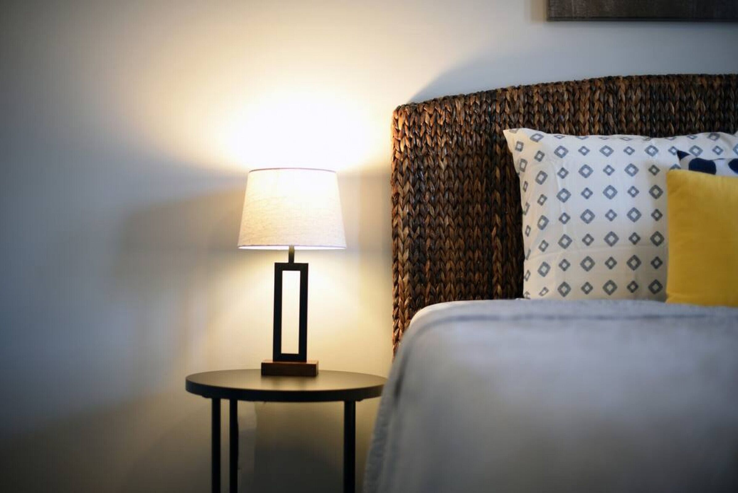 A neatly made bed with patterned pillows, a round bedside table with a lamp, and a textured wall.