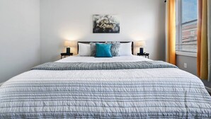 A bedroom with a large bed, two bedside tables with lamps, a wall-mounted artwork, and a window with blinds.