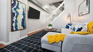 A modern living room with a grey sofa, a yellow throw blanket, a black and white patterned rug, a large abstract painting, and a staircase leading to another level.