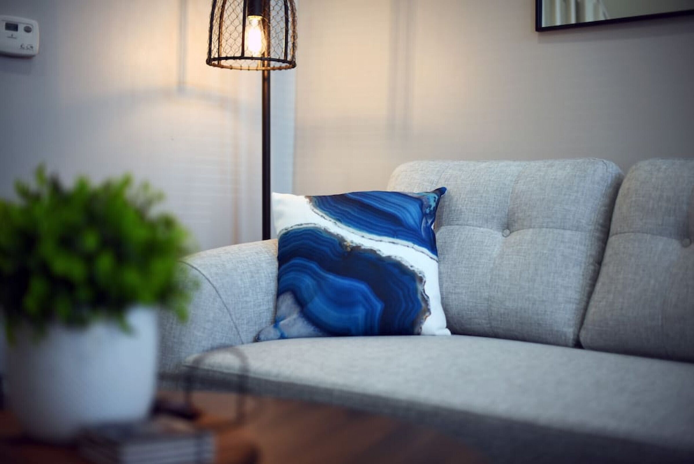 Un salón moderno con un sofá gris, un cojín azul y blanco estampado, una lámpara de pie y una planta en maceta.