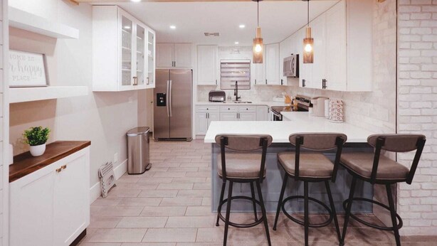 A modern kitchen with white cabinets, a stainless steel refrigerator, a central island with bar stools, and pendant lighting.