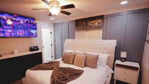 A bedroom with a tufted headboard, a flat-screen TV, a bedside table with a lamp, and a large window.