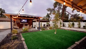 A backyard with artificial grass, a covered seating area, and string lights.