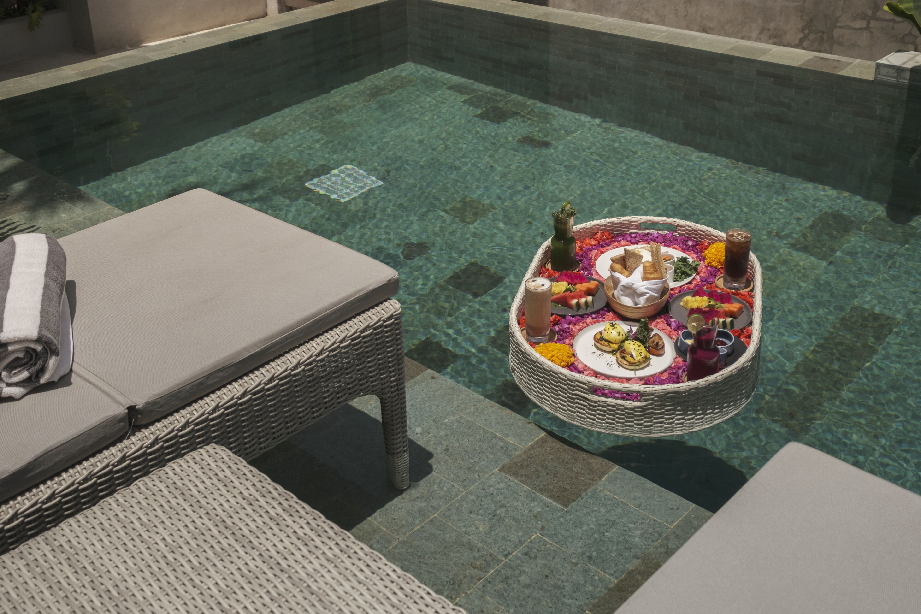 Zona de piscina con mesa de mimbre dispuesta con comida y bebidas, una silla de descanso y una mesita con botella y vasos.