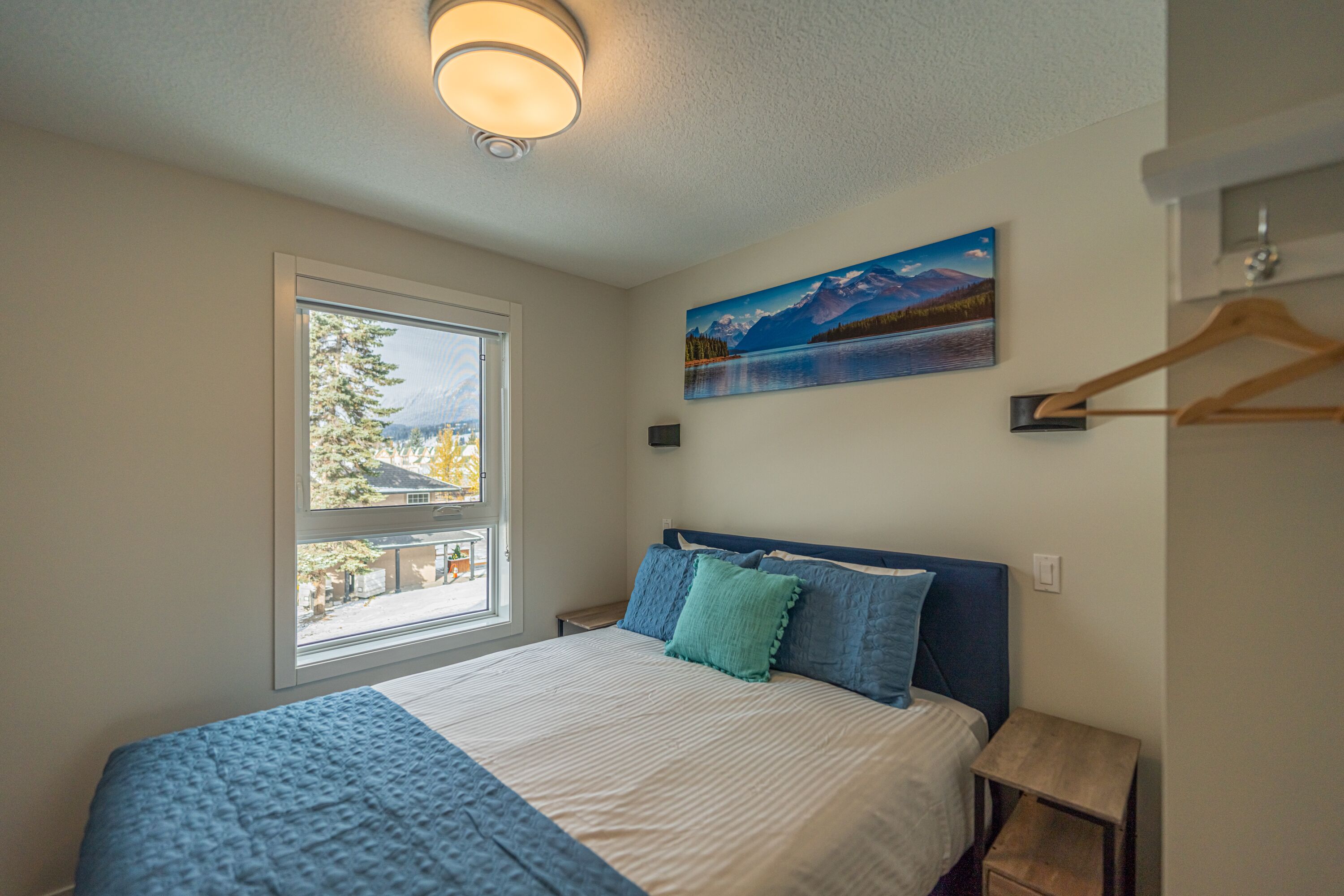 A bedroom with a bed, a window, a painting, and a wooden hanger.