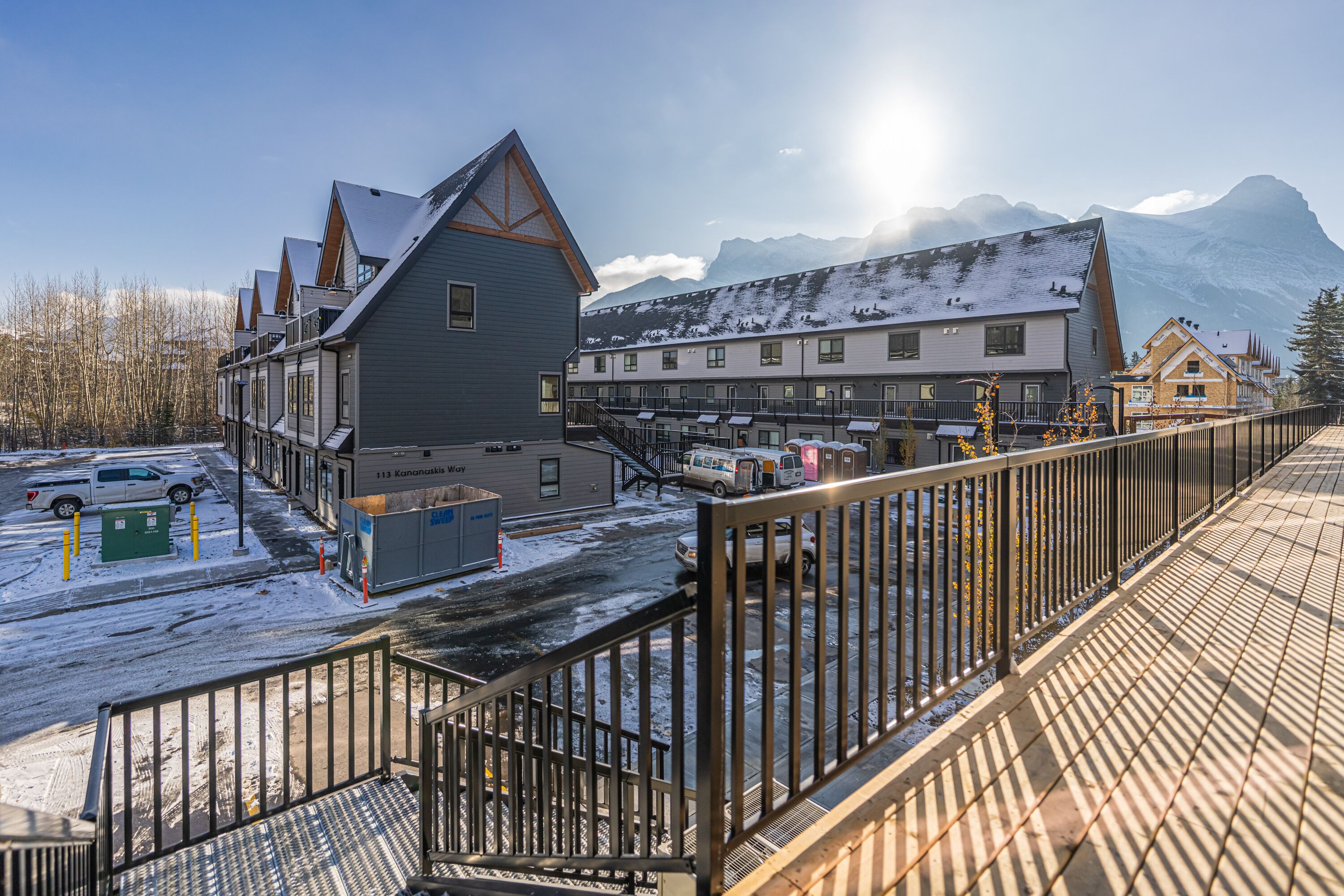 A hotel with a modern design, featuring a mix of residential and commercial buildings, a parking area, and a view of snow-covered mountains.