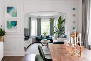 Relax in this stylish living room with a sofa, coffee table, and dining area, surrounded by abstract art and lush greenery.