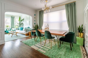 Et spiseområde med et træbord, grønne stole og en grøn måtte. Der er krukkeplanter og et stort vindue med gardiner.