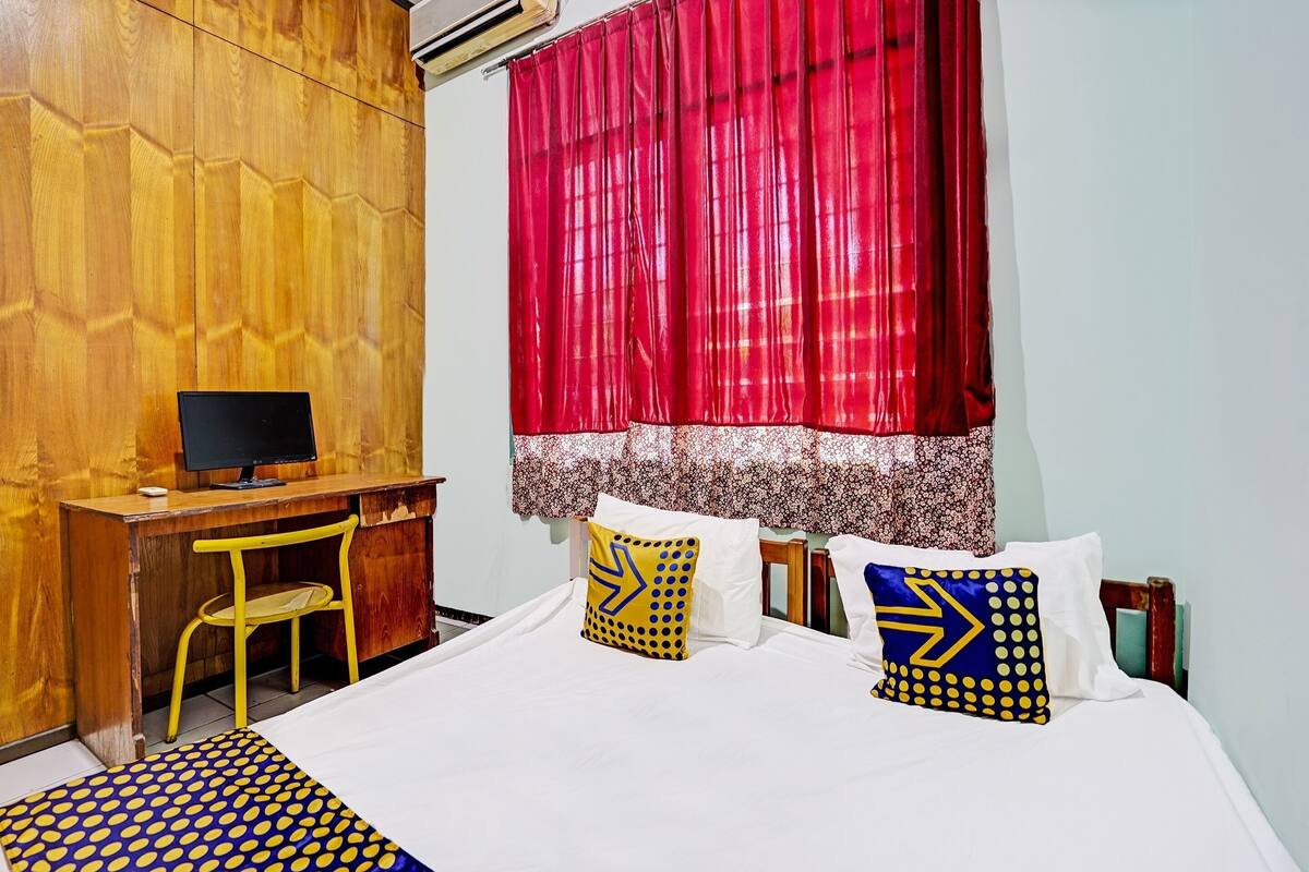 Un dormitorio con un escritorio de madera, una silla, una cama con almohadillas estampadas y una ventana con cortinas rojas.