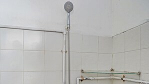 Un baño pequeño con azulejos, ducha de mano, repisa de vidrio y una tubería visible.
