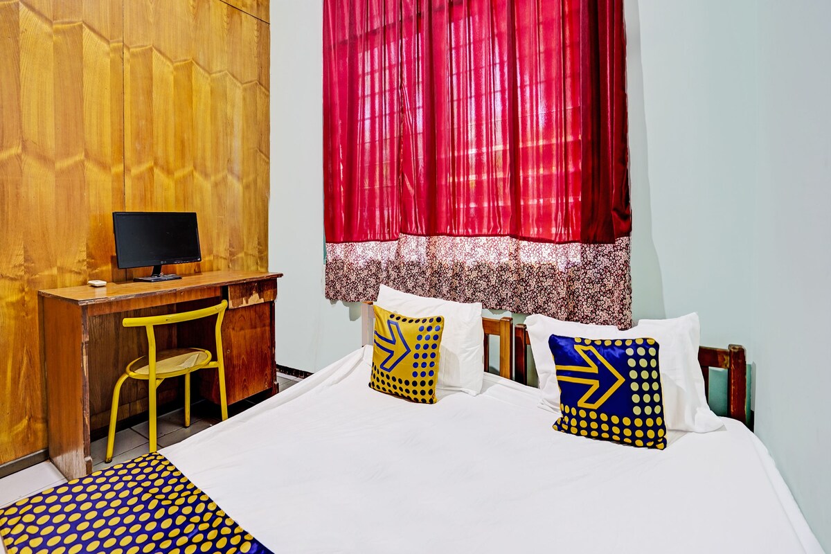 A bedroom with a wooden desk, a computer, a chair, a bed with patterned pillows, and a window with red curtains.