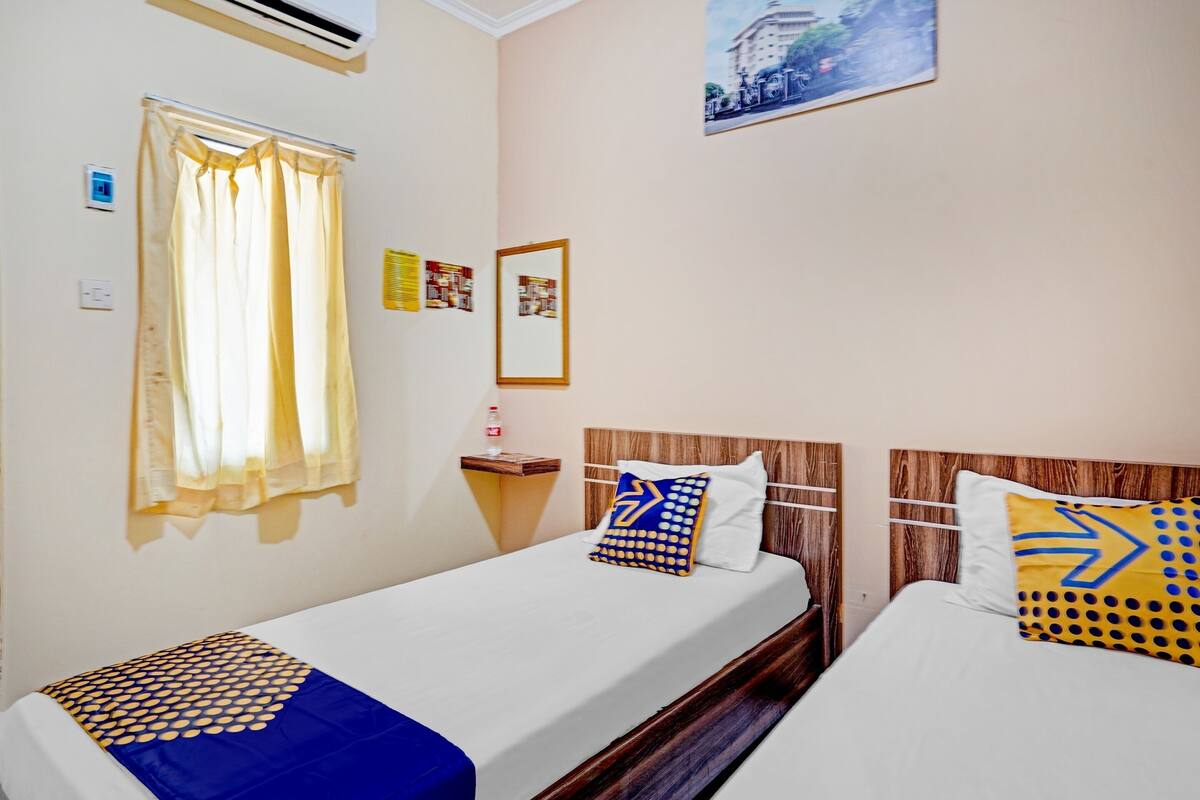 A hotel room with two single beds, a window with curtains, a wall-mounted air conditioner, a small table with a bottle, and a framed picture on the wall.