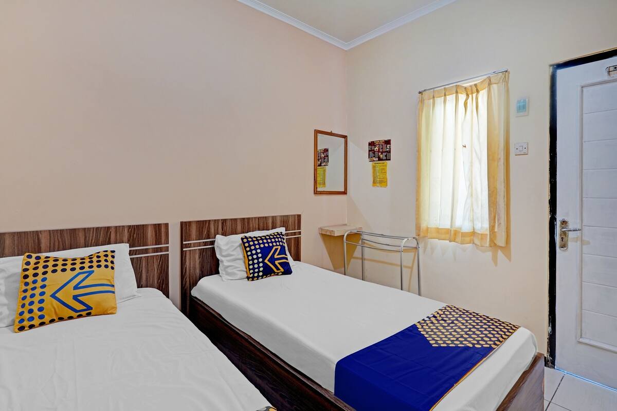 A hotel room with two beds, each with a different patterned pillow, a small table, a window with curtains, and a door.