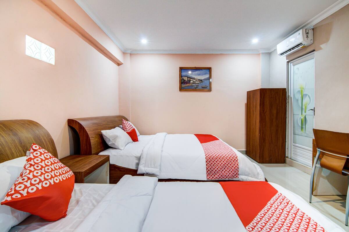 Une chambre d’hôtel avec deux lits, une tête de lit en bois, un oreiller à motifs rouges et blancs, une table de chevet en bois, une armoire en bois et un tableau encadré au mur.