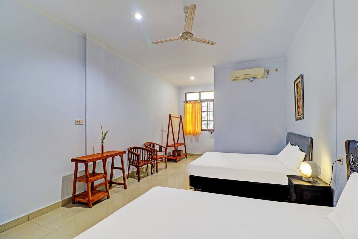 A hotel room with two beds, a desk, chairs, a ceiling fan, and a window with curtains.