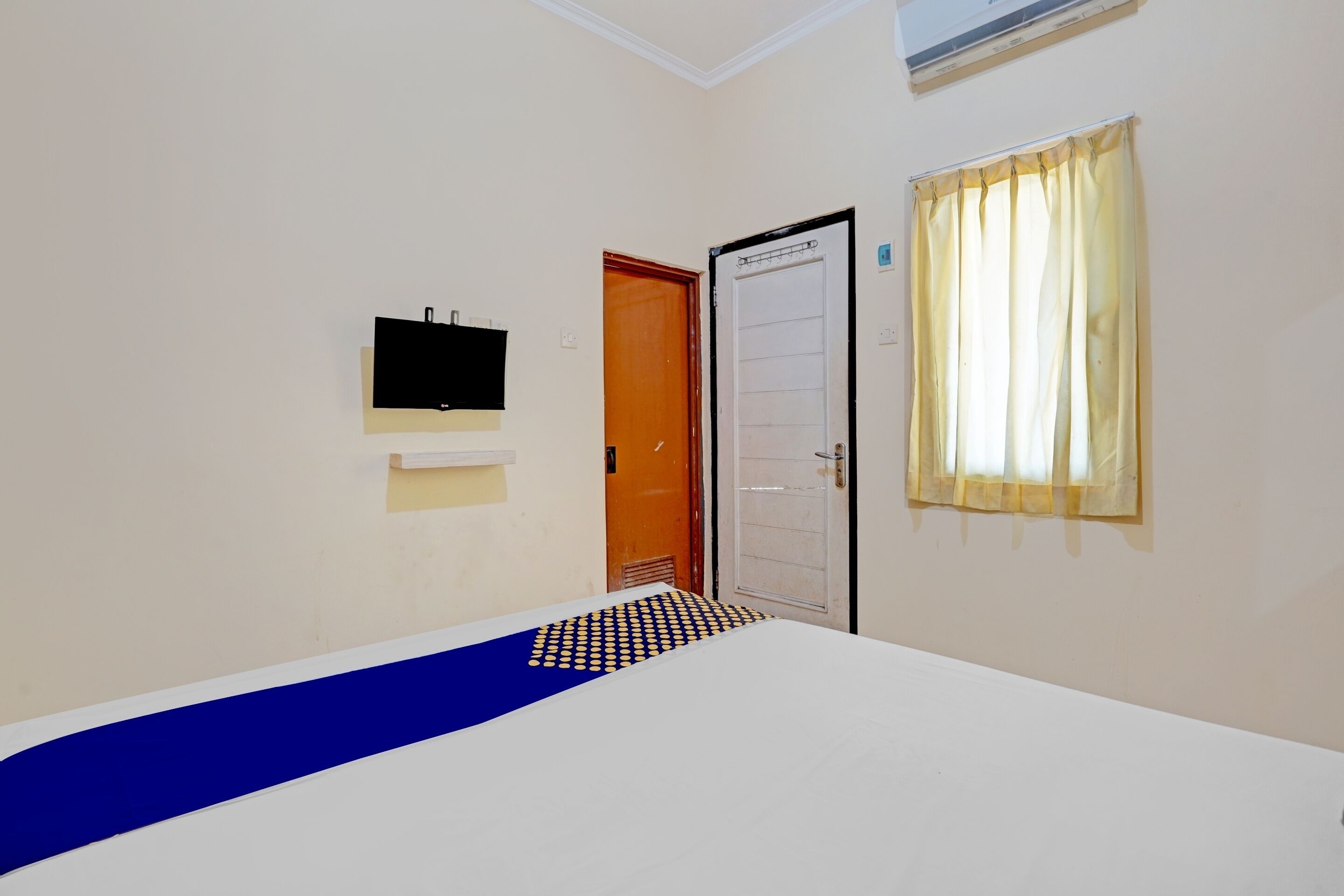 A room with a bed, a television mounted on the wall, a door, and a window with curtains.