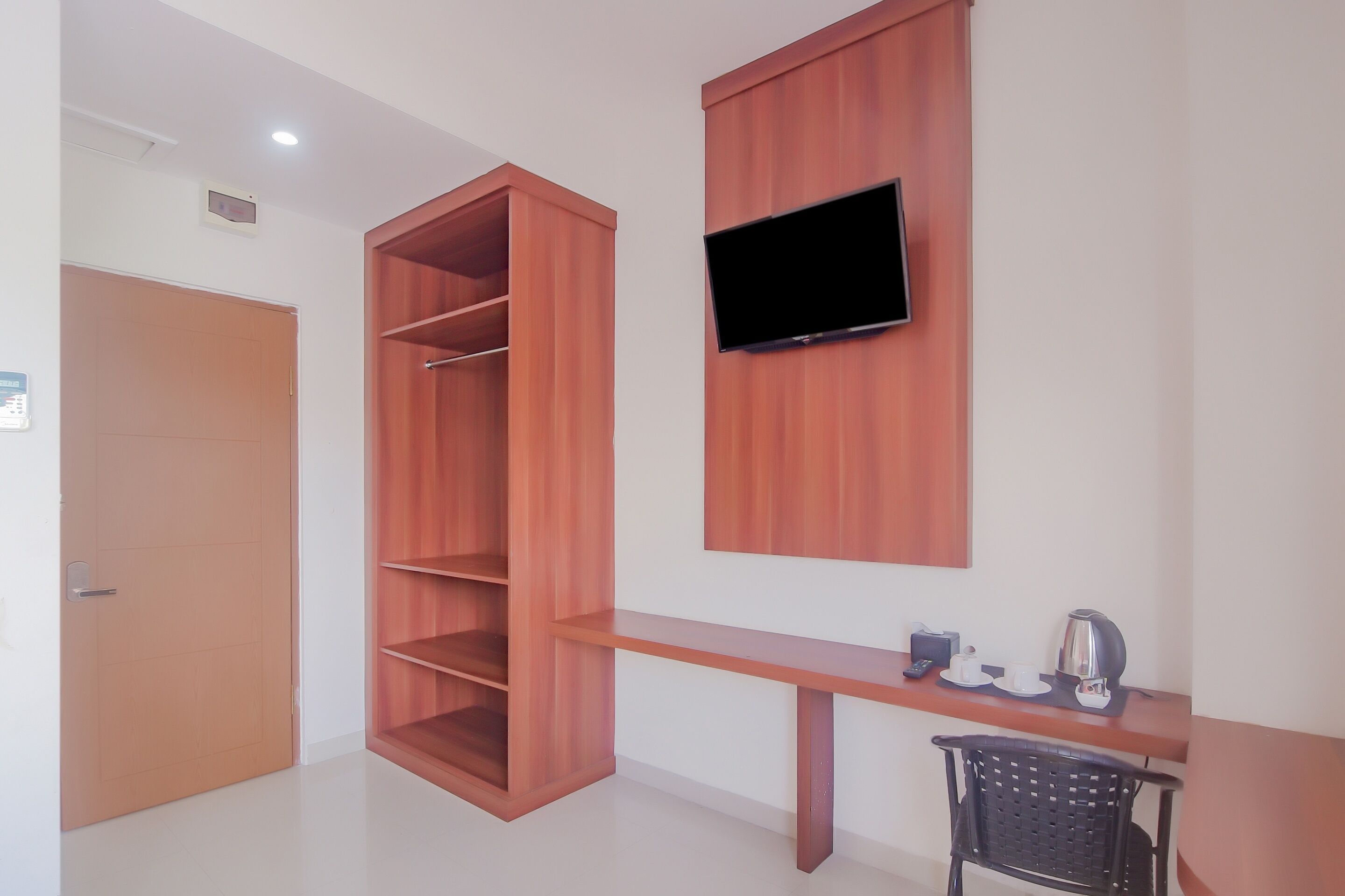 A room with a wooden wardrobe, a mounted TV, a desk with a chair, and a coffee maker.