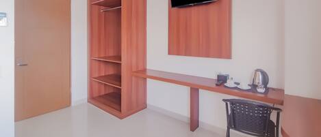 A room with a wooden wardrobe, a mounted TV, a desk with a chair, and a coffee maker.