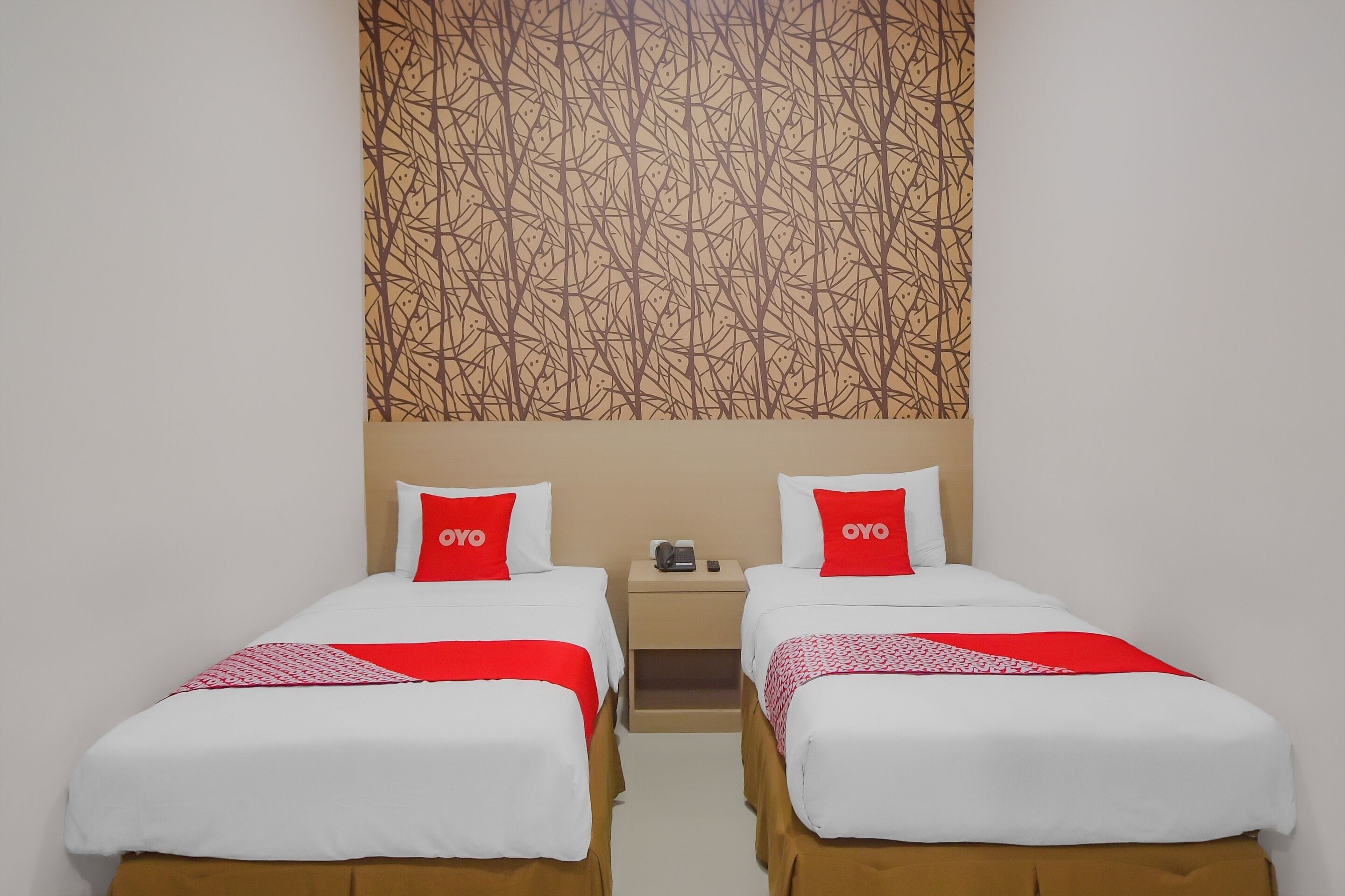 A hotel room with two single beds, each with a red and white bedspread, and a small bedside table with a phone and a cup.