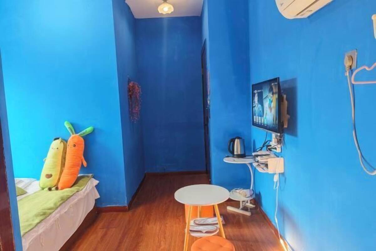 Ein kleines Zimmer mit blauen Wänden, einem Bett mit grüner Bettdecke, einem kleinen Tisch mit Stuhl, einem an der Wand befestigten Fernseher und einem Wasserkocher auf einem kleinen Tisch.