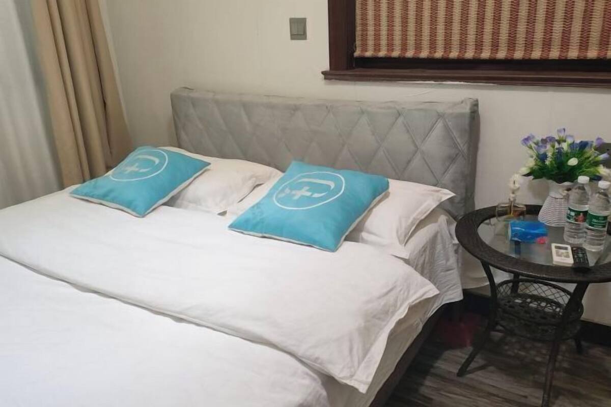 Ein Hotelzimmer mit einem Bett, zwei Kissen, einem kleinen Tisch mit Blumenvase und einem Fenster mit Vorhängen.