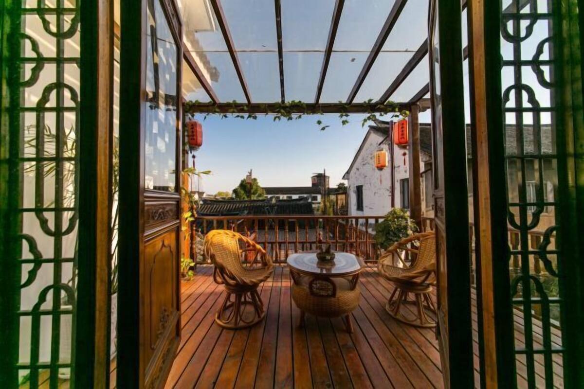Eine Holzterrasse mit Korbmöbeln und einer Glaskuppel, mit Blick auf ein traditionelles Gebäude mit roten Laternen.