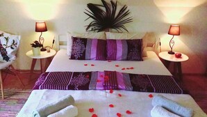 Habitación de hotel con una cama bien hecha, dos lámparas de noche, una planta decorativa y una mesita con fruta.