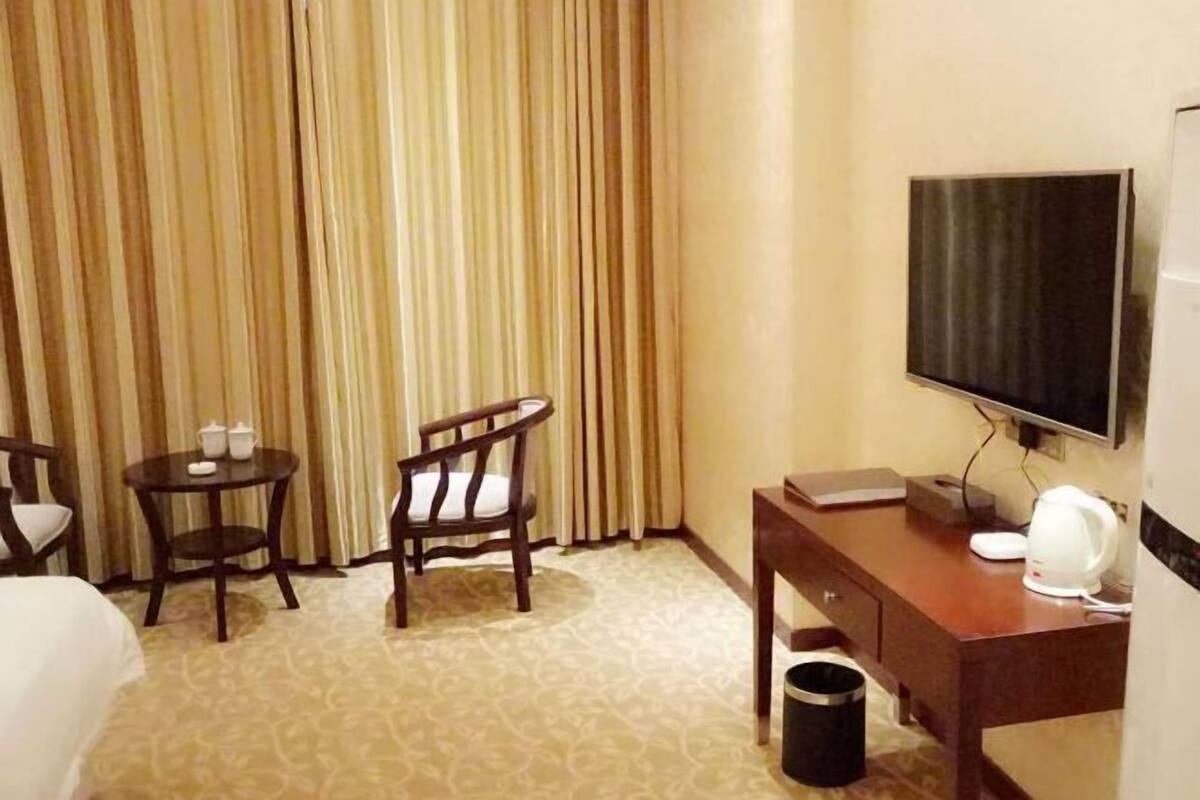 A hotel room with a bed, two chairs, a small table with a kettle, a TV, and a trash bin.