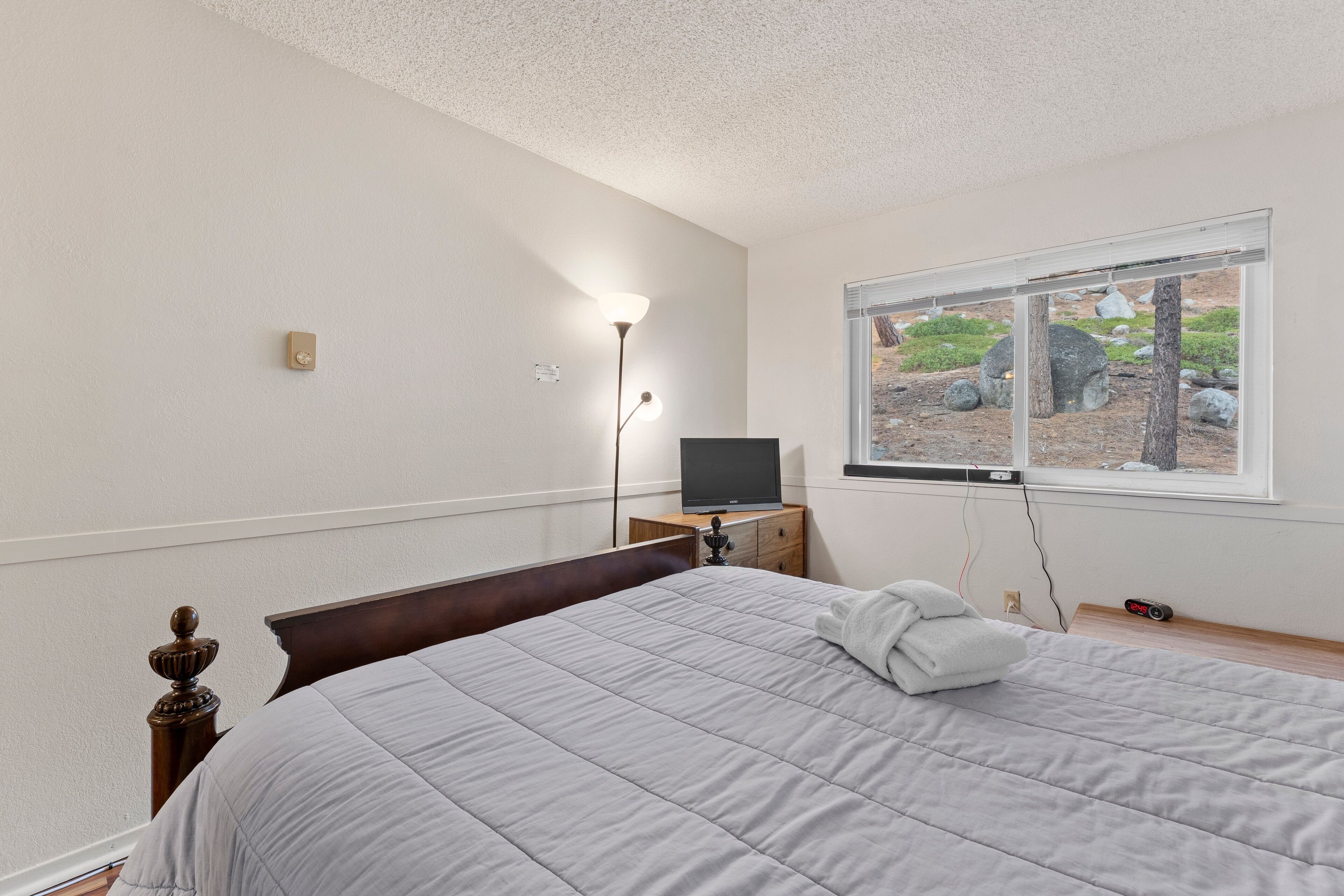 Une chambre à coucher avec un lit, un téléviseur posé sur une commode en bois, une lampe et une fenêtre donnant sur des rochers et de la verdure.
