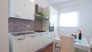 Eine moderne Küche mit weißen Schränken, einer Edelstahl-Spüle und einem eingebauten Backofen. Ein Esstisch mit zwei Stühlen und einer Weinflasche steht daneben.