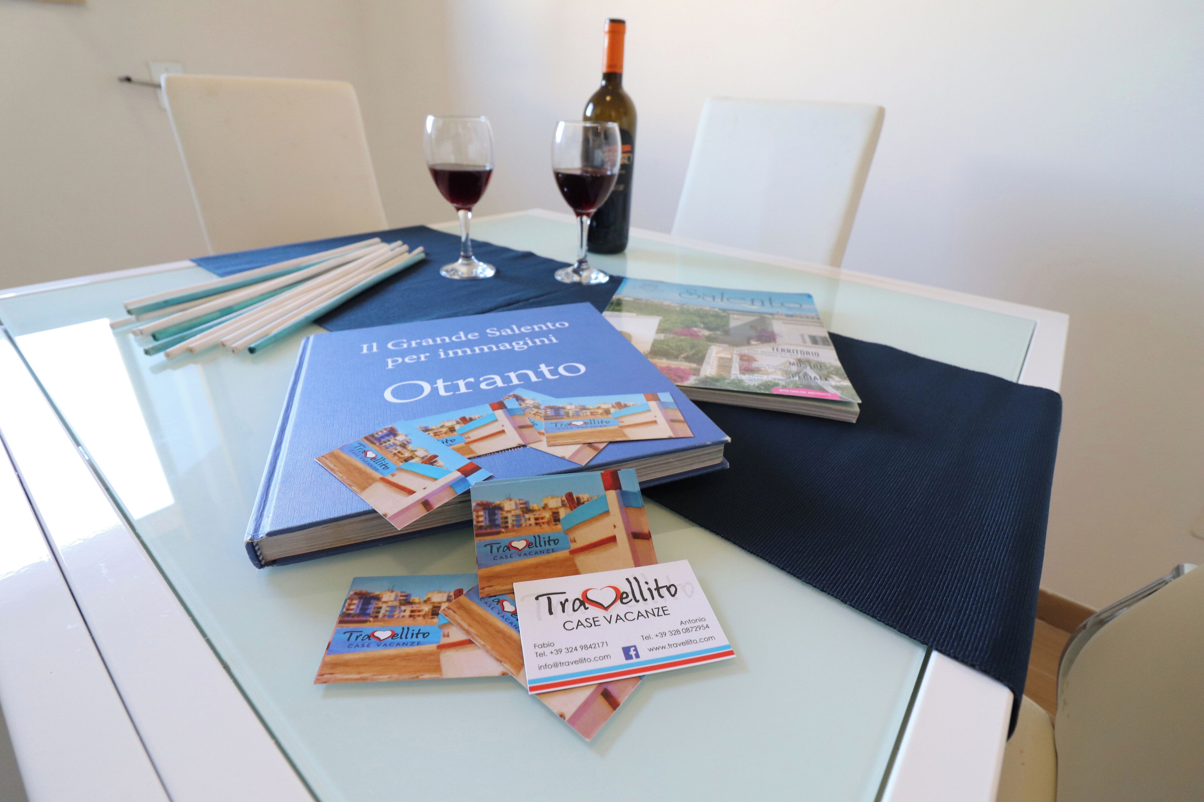 A glass table with a book titled 'Il Grande Salento per Immagini Otranto', a bottle of wine, and business cards for 'Tra'ellitto'.