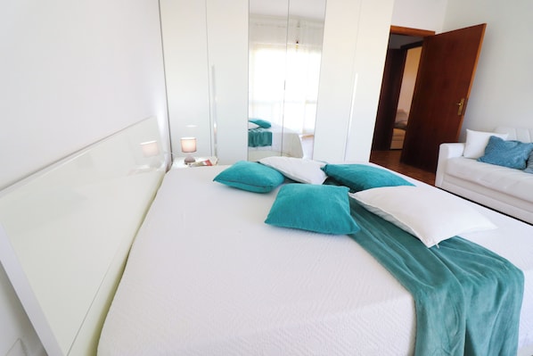 A bedroom with a bed, pillows, a bedside table, a mirror, and a white sofa.