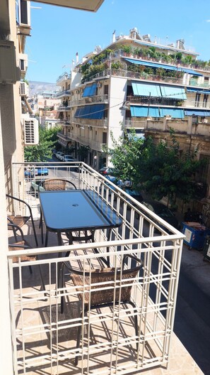 Un balcón con mesa y sillas, con vistas a una calle con coches aparcados y edificios.