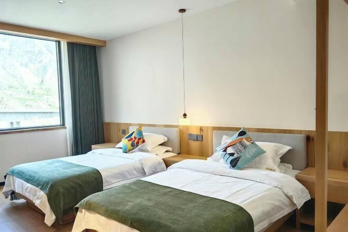 A hotel room with two beds, a window with curtains, a hanging light fixture, and a wooden headboard.