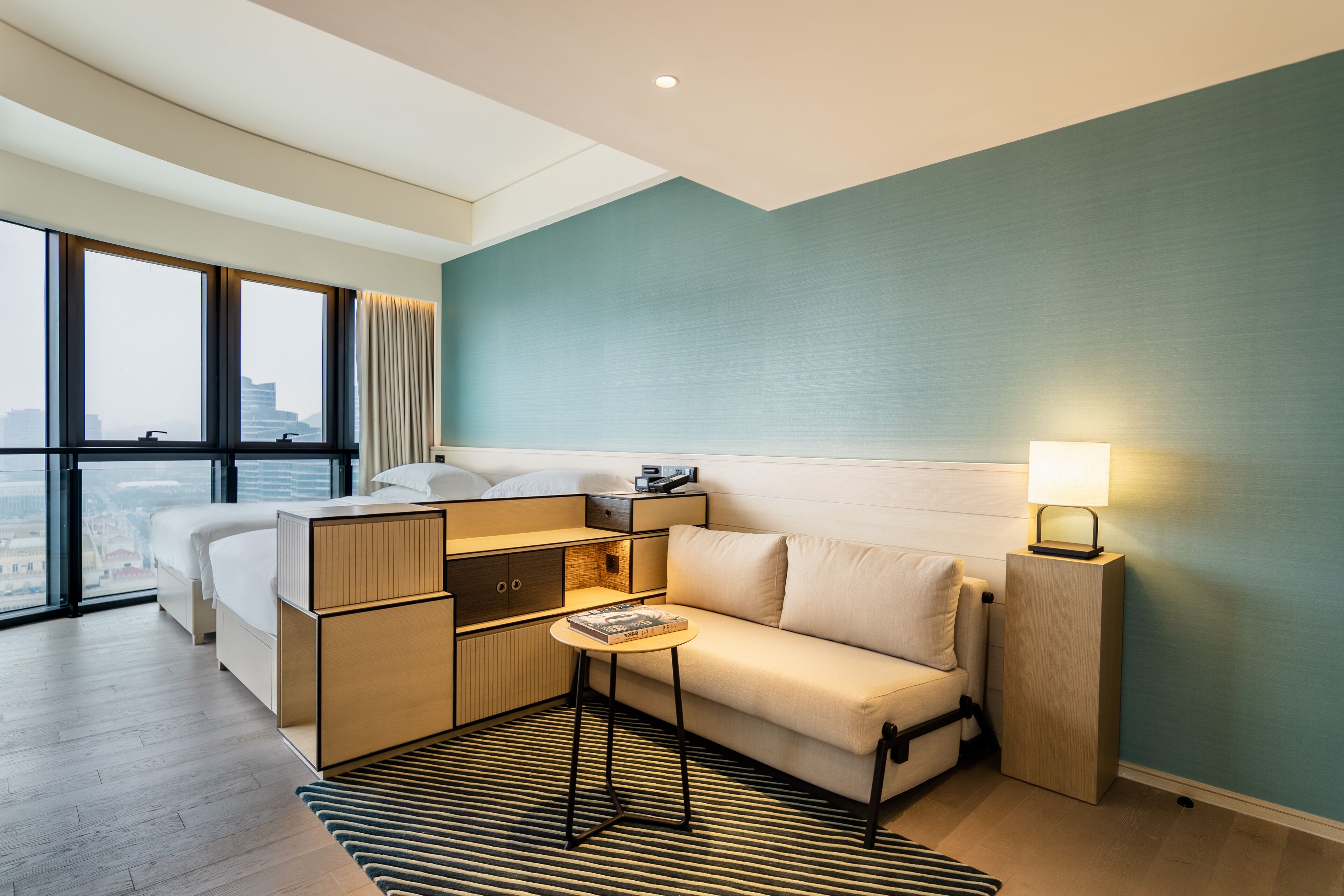 Une chambre d’hôtel moderne, équipée d’un lit, d’un canapé, d’une petite table et d’une table de chevet. Une grande fenêtre offre une vue sur la ville.