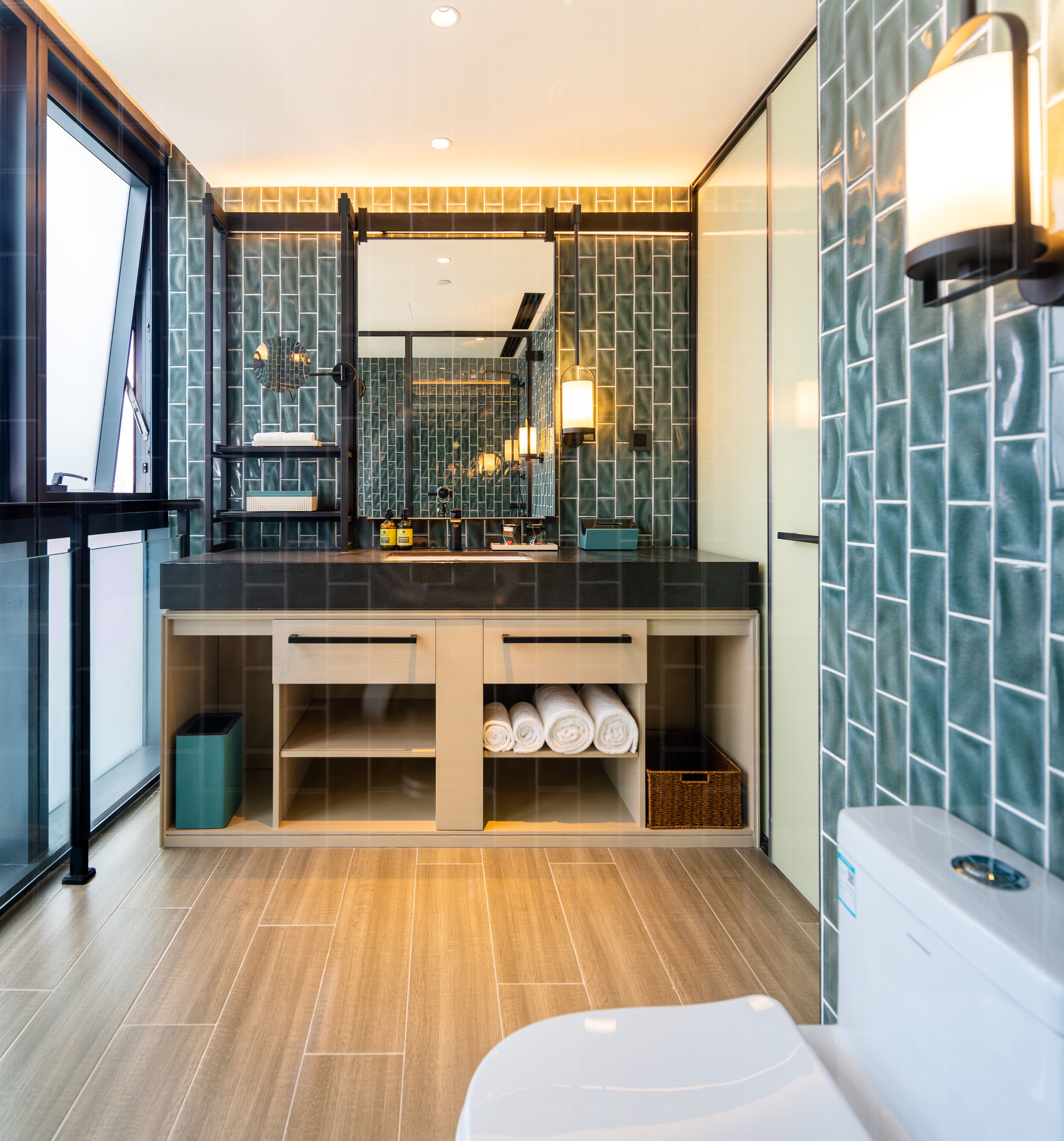 Un baño moderno con un espejo grande, paredes de azulejos oscuros, suelo de madera y almacenamiento integrado.