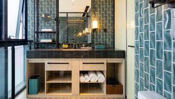 Un baño moderno con un espejo grande, paredes de azulejos oscuros, suelo de madera y almacenamiento integrado.