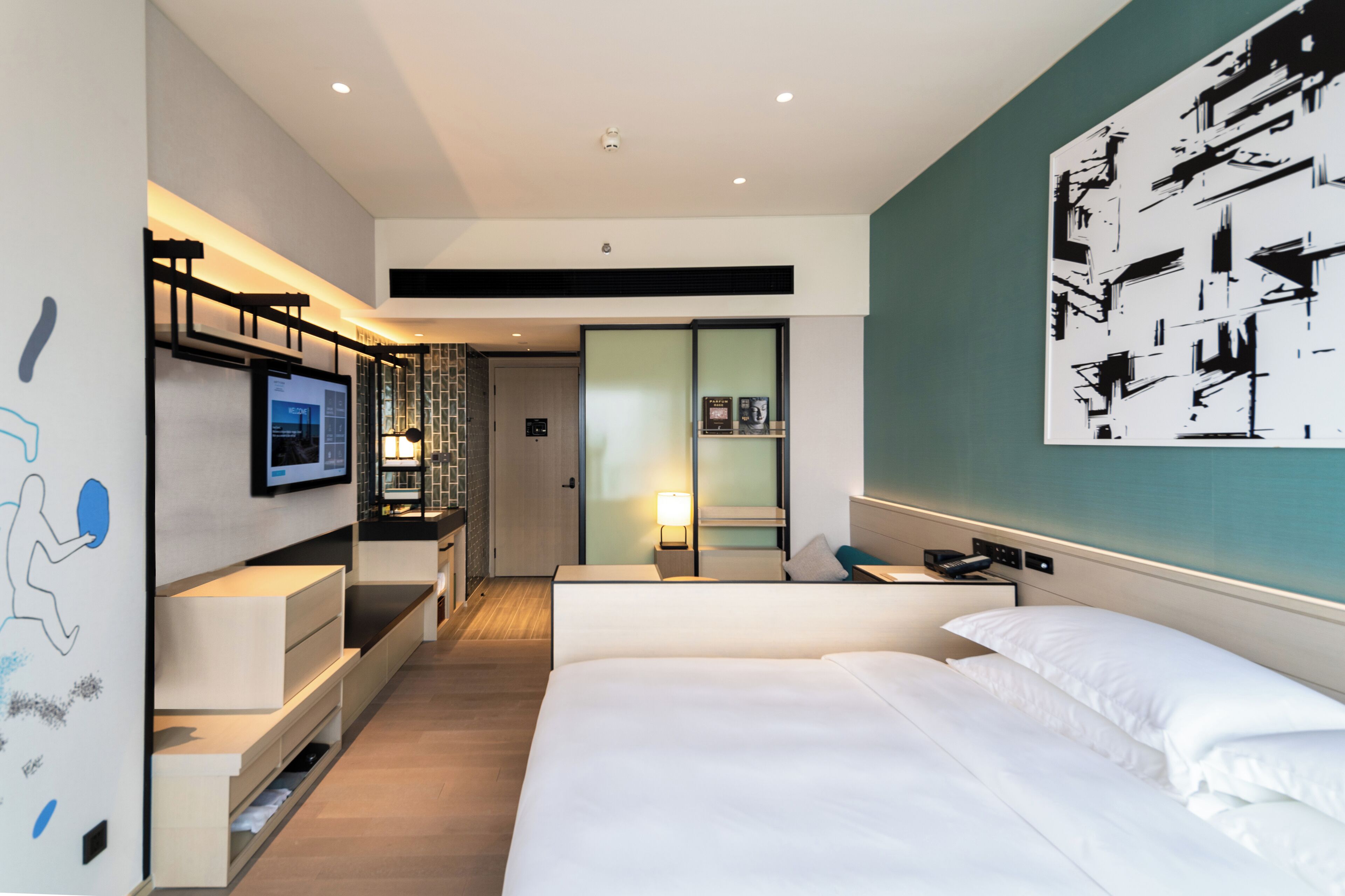 A modern hotel room with a large bed, a TV mounted on the wall, a desk with a chair, and a bathroom visible through a glass door.