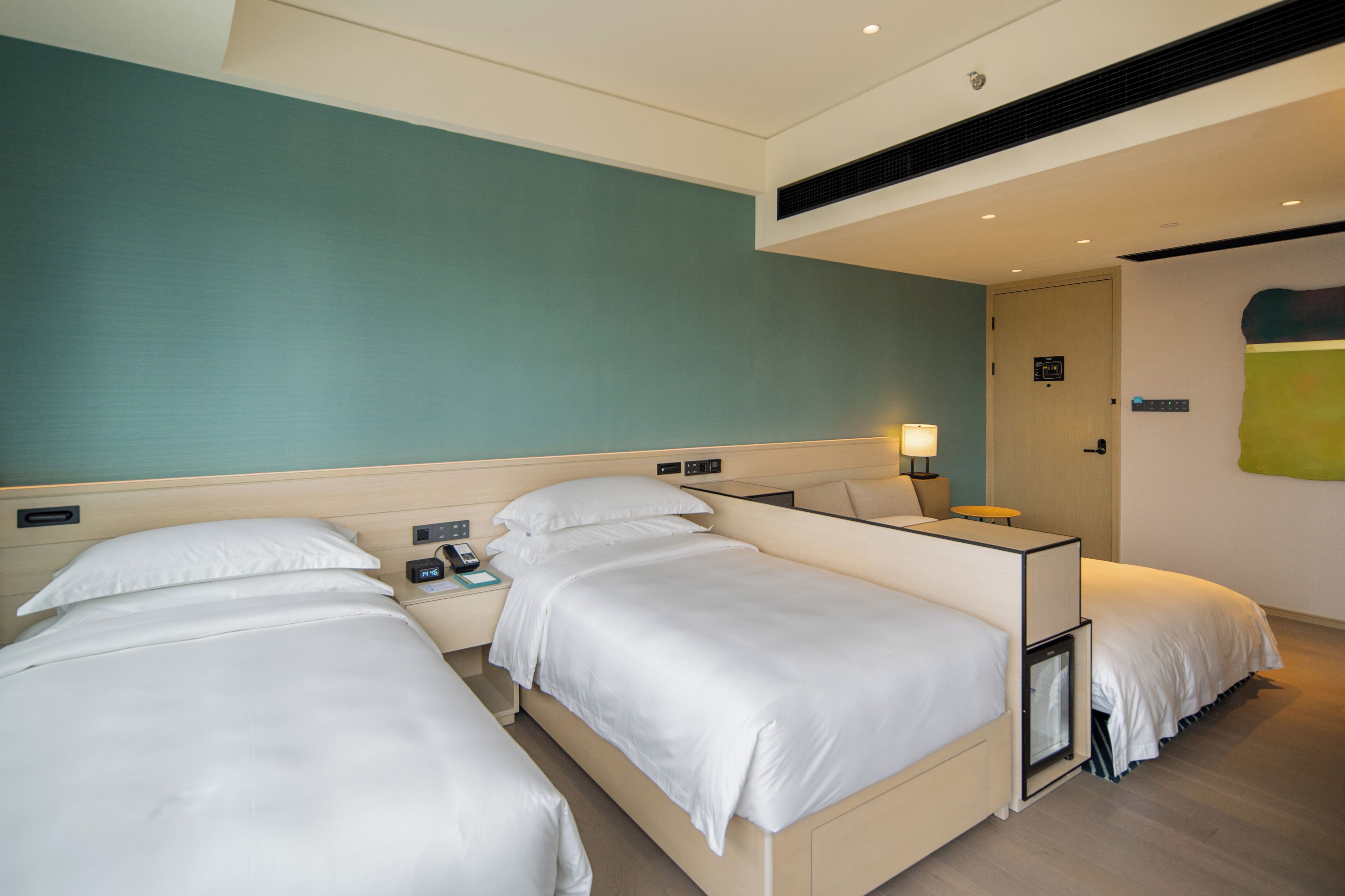 A hotel room with two beds, a bedside table with a lamp, and a small bedside table with a phone and a book.