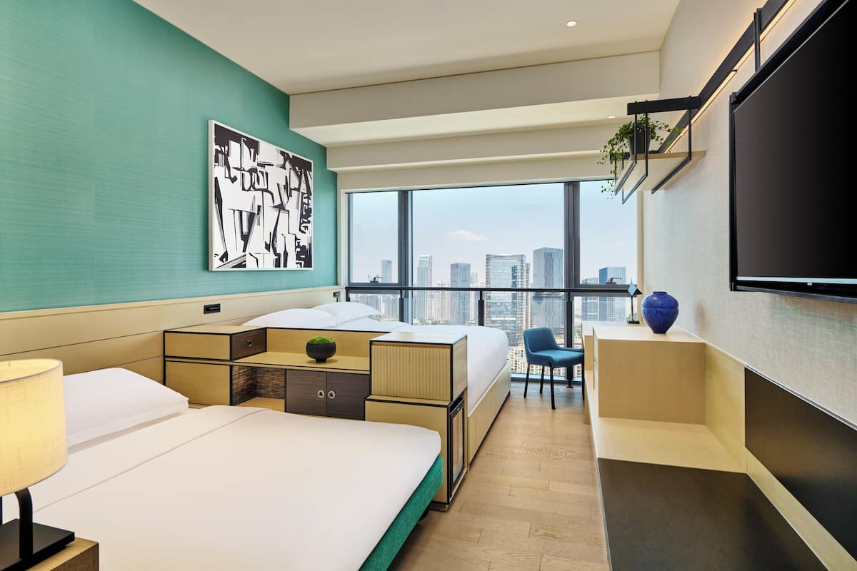 A hotel room with two beds, a large window with a city view, a mounted TV, and a small desk with a plant.