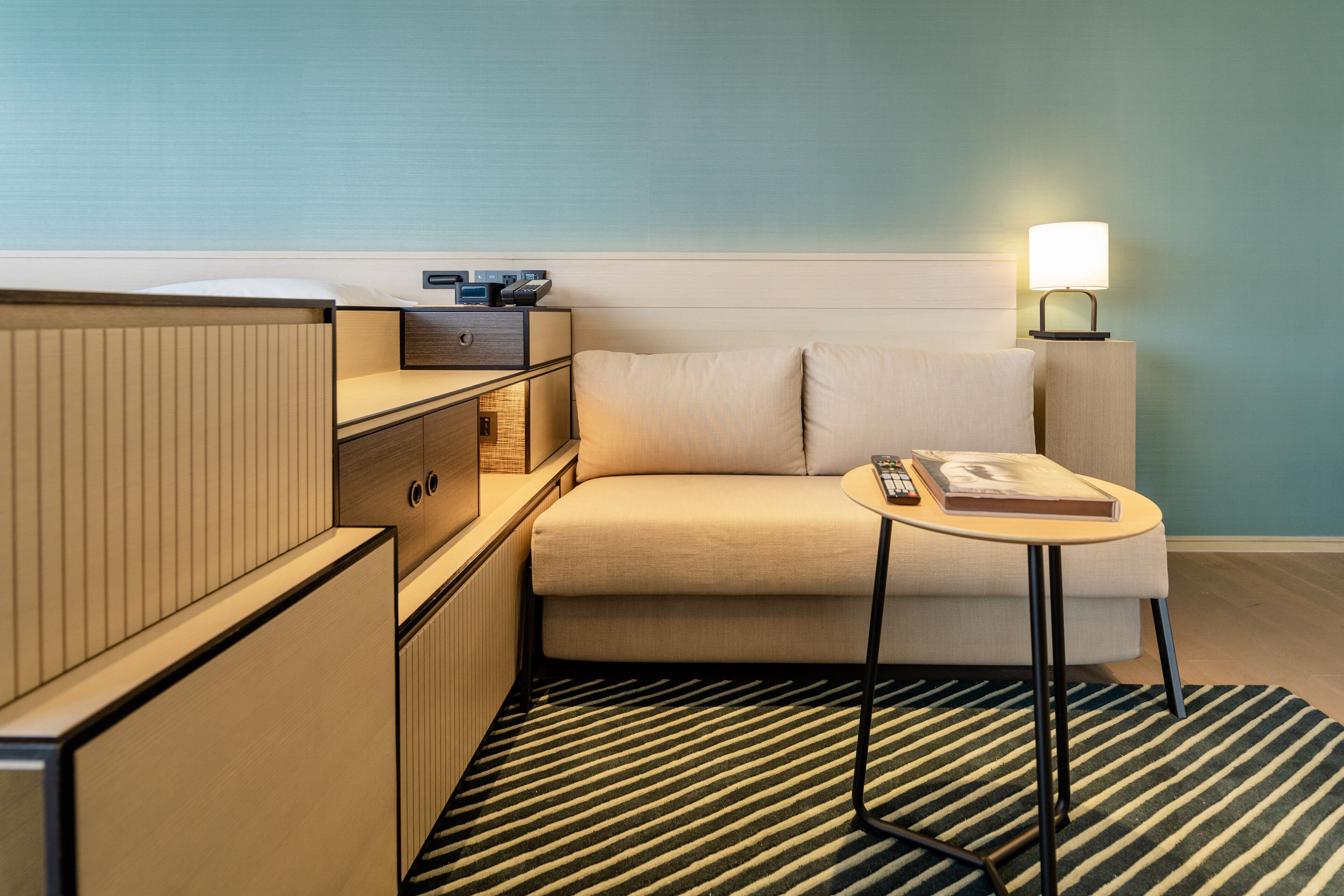 A hotel room with a sofa, a small table with a lamp, a remote control, and a book.