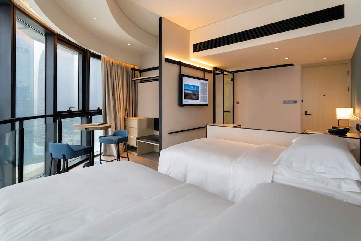 A hotel room with two beds, a desk, a chair, a large window with curtains, and a mounted digital screen.
