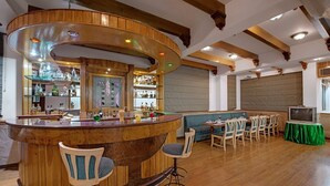 A bar area with a curved wooden counter, multiple stools, and a television on a table. - Hotel Baljees Regency (Shimla)