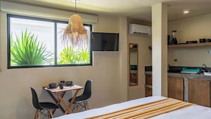 Une chambre d’hôtel comprenant un lit, un bureau avec des chaises, une télévision, un coin cuisine et une grande fenêtre avec des plantes.