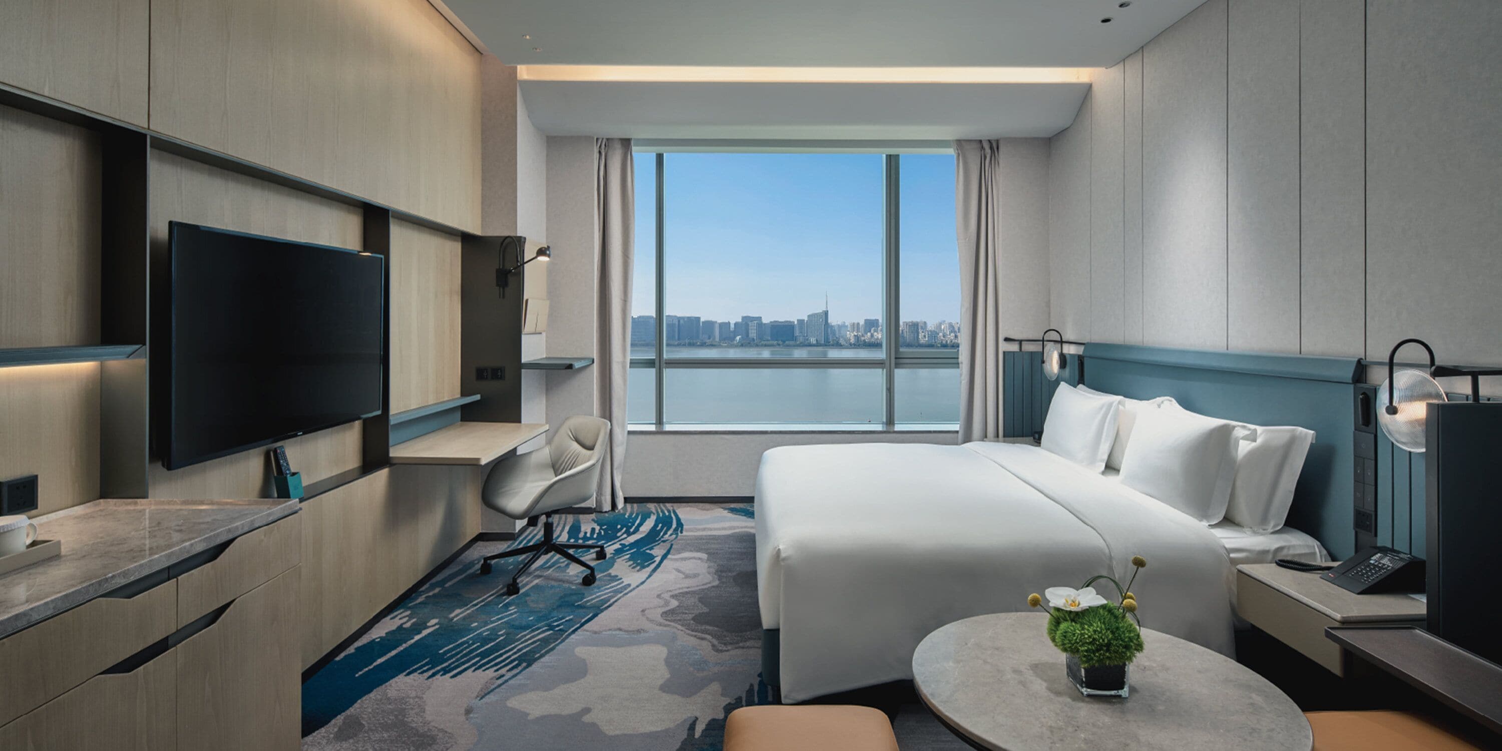 A modern hotel room with a large bed, a desk, a flat-screen TV, and a view of a city skyline.