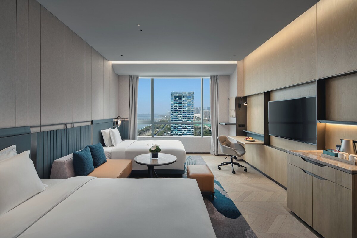 Quarto de hotel moderno com cama grande, escrivaninha, poltrona, televisão e vista para a paisagem urbana.