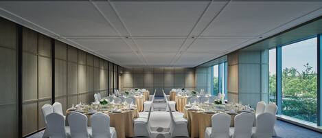 Sala de conferência com mesas redondas, cadeiras brancas e amplas janelas com vista para o verde.