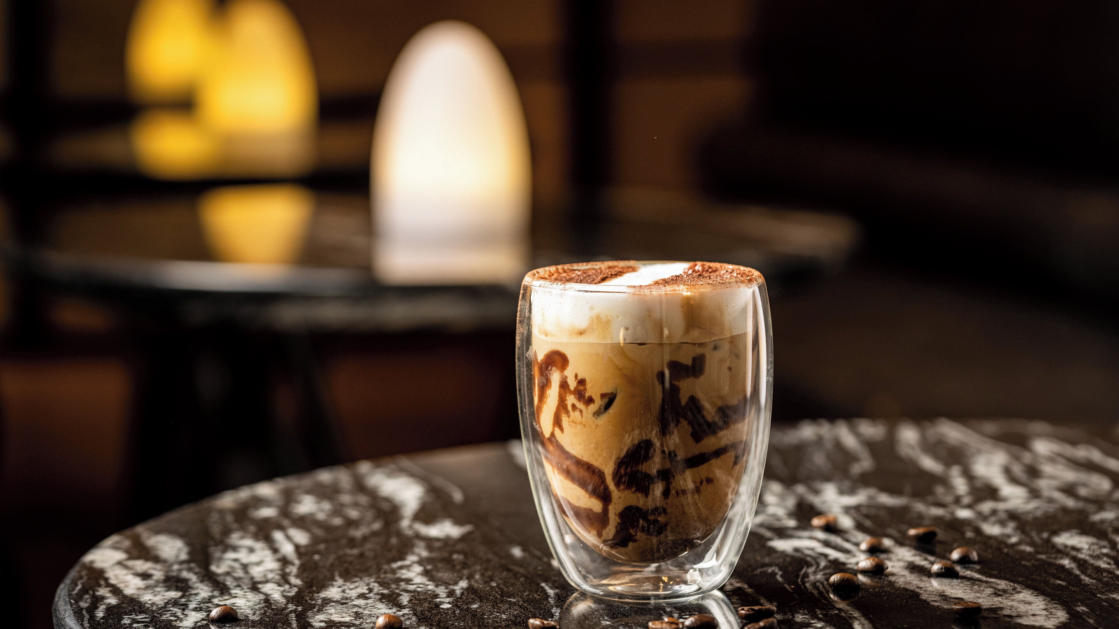 Ein Glas mit einem schichtartigen Getränk und schaumiger Oberfläche, auf einer strukturierten Fläche mit verstreuten Nüssen platziert.