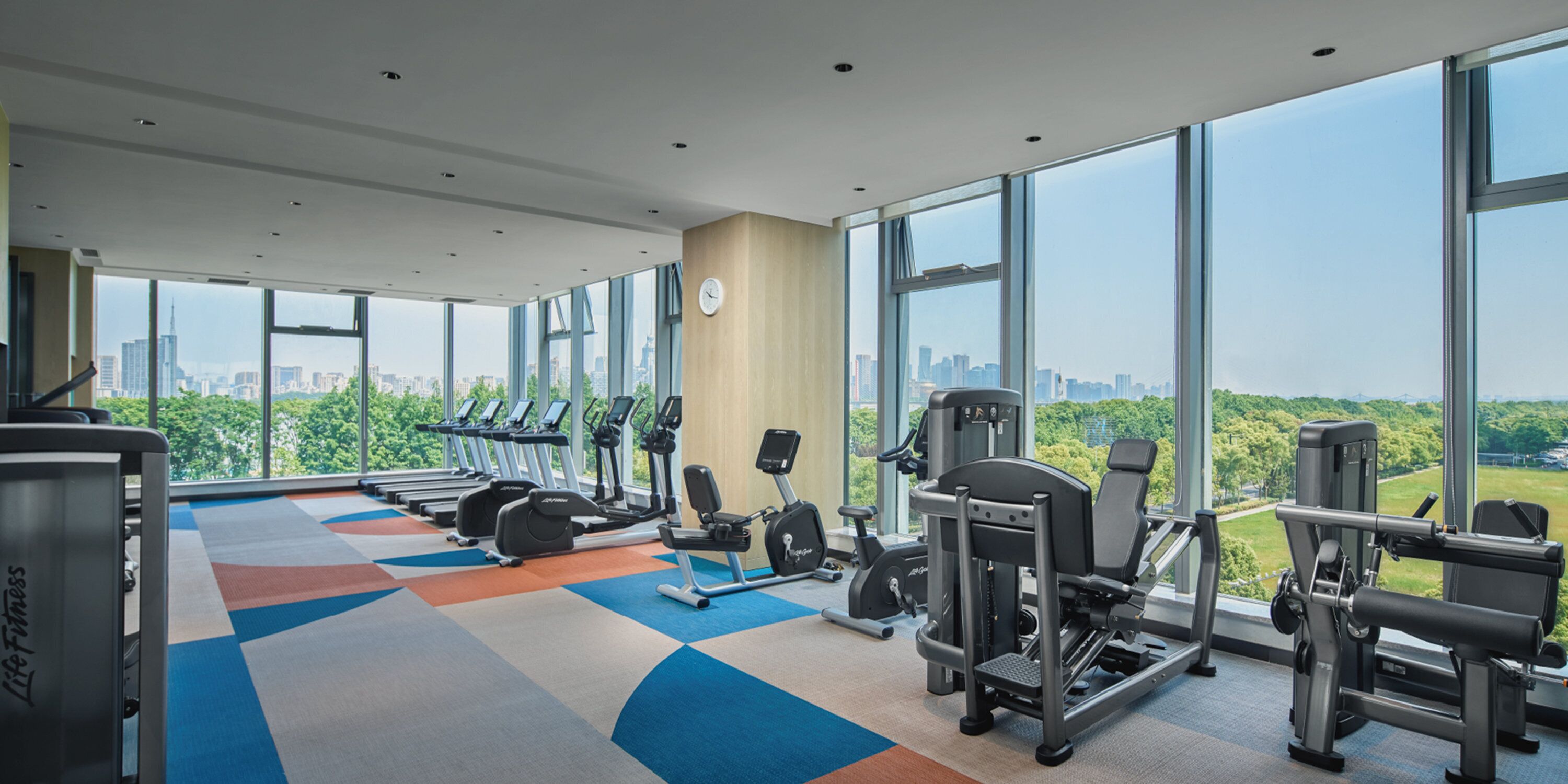 Ein Fitnessstudio mit Cardiogeräten, Krafttrainingsgeräten und einem großen Fenster mit Blick auf die Stadt.