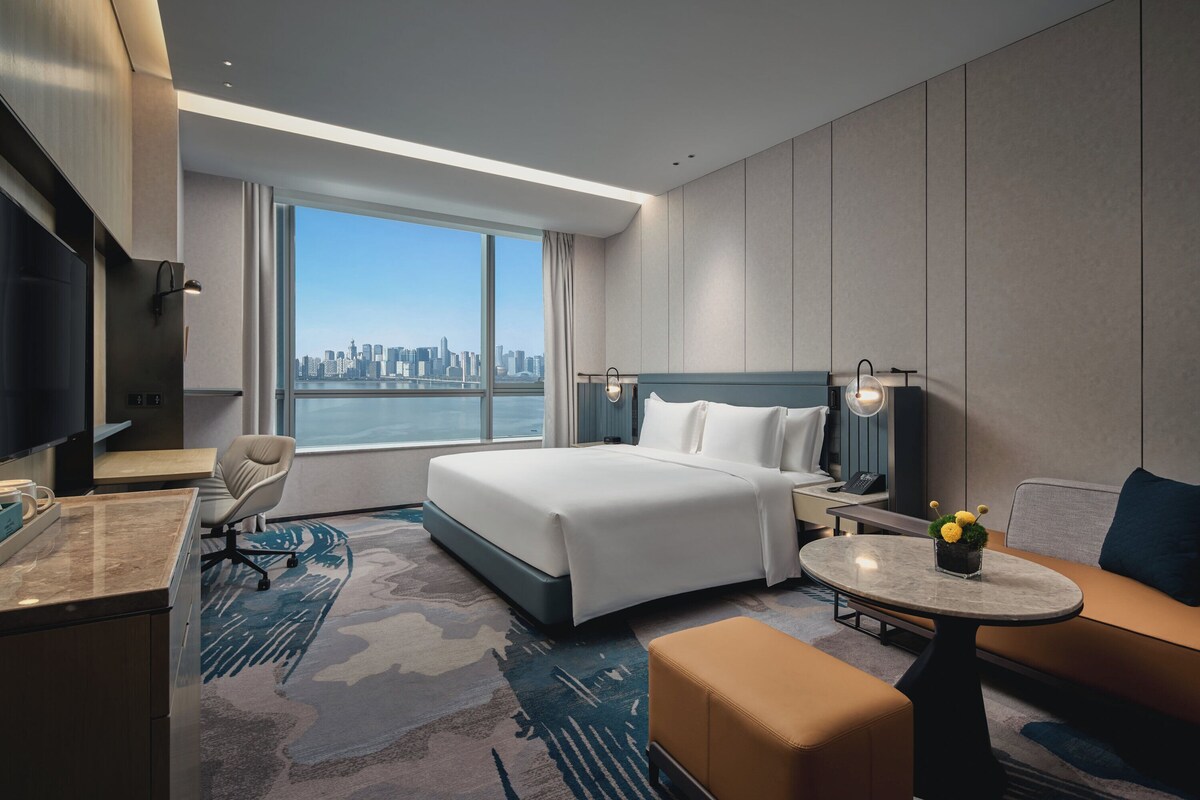 Quarto de hotel com uma cama grande, uma escrivaninha, uma cadeira, um sofá e vista para o horizonte da cidade.
